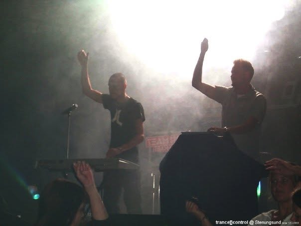 The two members on a smoky stage with their arms raised, behind a keyboard and a covered rack.